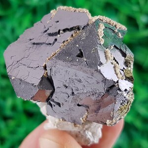Puede incluir: Un cristal met&aacute;lico gris oscuro con una superficie rugosa y texturizada. El cristal tiene una forma geom&eacute;trica con m&uacute;ltiples facetas. Est&aacute; rodeado de una sustancia blanca y pulverulenta.
