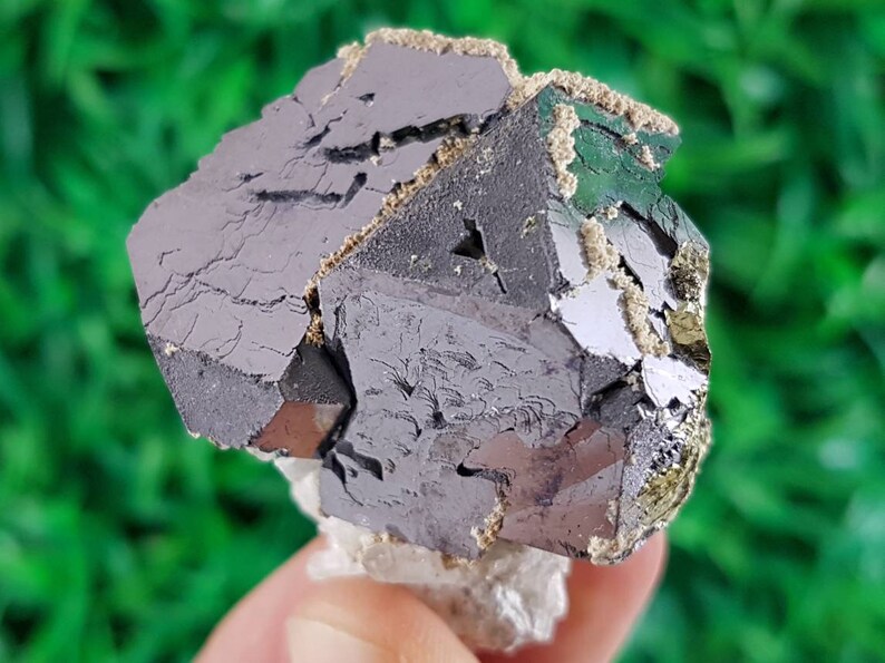 Puede incluir: Un cristal met&aacute;lico gris oscuro con una superficie rugosa y texturizada. El cristal tiene una forma geom&eacute;trica con m&uacute;ltiples facetas. Est&aacute; rodeado de una sustancia blanca y pulverulenta.