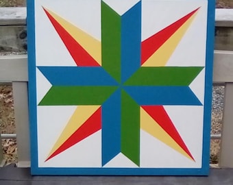 May include: A colorful barn quilt with a star design. The star is made up of red, yellow, green, and blue triangles. The quilt is painted on a white wooden board with a blue border.
