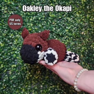 May include: A handmade crochet okapi plush toy. The okapi has a brown body, a black and white patterned tail, and a black face. The text "Oakley the Okapi" is displayed at the top of the image.