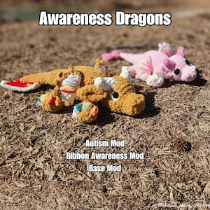 May include: Two handmade crochet dragons in a natural outdoor setting. One is tan with rainbow accents, the other is pink. Text at the top reads "Awareness Dragons." Additional text includes "Autism Mod," "Ribbon Awareness Mod," and "Base Mod."