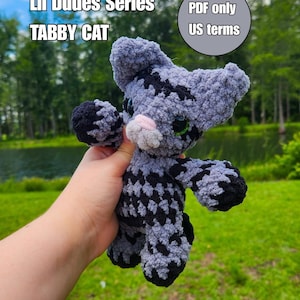 Op de afbeelding: Een gehaakt grijs en zwart cyperse kat knuffeltje met grote groene ogen en een roze neus. Tekst op de afbeelding luidt: Lil Dudes Series TABBY CAT en PDF only US terms. De knuffel heeft een getextureerd, pluizig garen uiterlijk met donkere strepen.