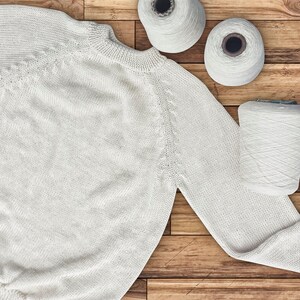 Puede incluir: Un suéter de punto blanco colocado plano sobre una superficie de madera con cuatro carretes de hilo blanco.