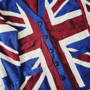 Puede incluir: Un cárdigan de punto con el diseño de la bandera de la Union Jack en rojo, blanco y azul. El suéter con botones tiene cuello en V y dos bolsillos delanteros. El diseño de la bandera se muestra en la parte delantera y las mangas.