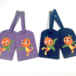 May include: Two luggage tags featuring a cartoon orange bird with a green leaf. One tag is purple and the other is navy blue.