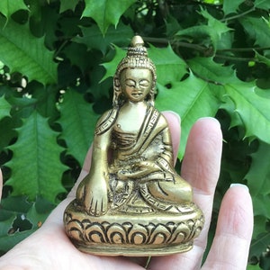 May include: A small gold-colored brass statue of Buddha sitting in meditation pose. The statue is detailed with intricate carvings and is sitting on a lotus base.