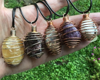 May include: Five pendants featuring polished, irregularly shaped stones encased in spiraled copper wire cages. Each stone displays a unique color, including yellow, dark brown, mottled tan, purple, and white with black specks, all suspended on black cords.