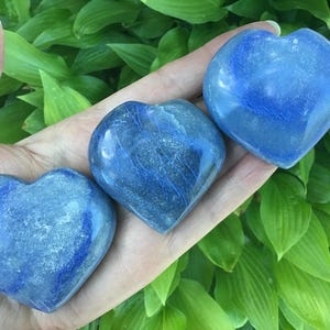 May include: Three heart-shaped blue stones with varying shades of blue and white marbling are displayed. The stones are held in a hand, with green foliage in the background. The stones are likely decorative or used for meditation.