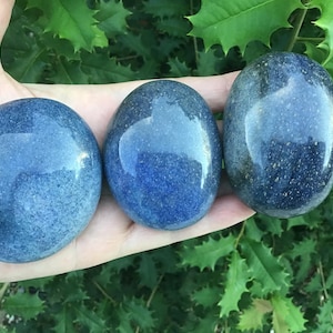 May include: Three smooth, polished blue aventurine stones with a rounded, oval shape. Each stone displays a deep, speckled blue color with subtle variations in tone and a glossy, reflective surface texture, showcasing natural mineral inclusions.