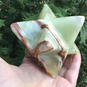 May include: A polished, multi-pointed star-shaped sculpture carved from light green onyx stone. The surface features smooth, swirling bands of brown and cream mineral inclusions that create a natural, marbled pattern across the geometric facets.