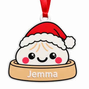 May include: A white dumpling ornament wearing a red Santa hat with a white pom-pom and trim. The dumpling has black eyes, a smiling mouth, and pink cheeks. It sits in a tan bamboo steamer base with the name Jemma printed in white letters.