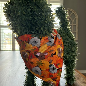 Puede incluir: Un perro de topiario artificial verde que lleva una bandana con un estampado de otoño de calabazas y perros. La bandana es naranja, marrón y blanca con un contorno negro.