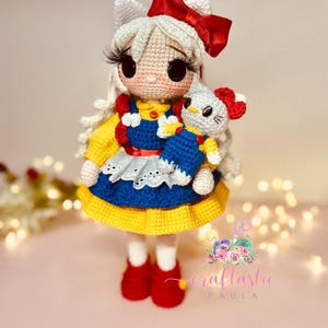 May include: A crocheted doll with white hair, a red bow, and a blue and yellow dress. The doll is holding a crocheted white kitten with red ears. The doll is standing on a pink surface.