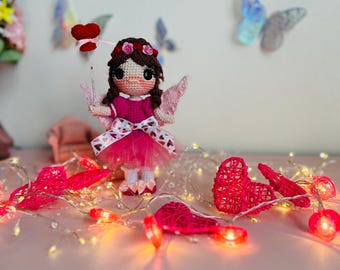 Puede incluir: Una muñeca de crochet con un vestido rosa y alas, sosteniendo una varita en forma de corazón rojo. La muñeca está rodeada de decoraciones en forma de corazón rosa y luces de cadena.