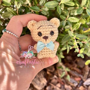 May include: A small, crocheted teddy bear in tan yarn with black safety eyes and a light blue bow tie. The bear is held in front of a green leafy background. The text "craftastic PAULA" is visible in the image.