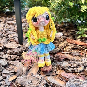 May include: A handmade crochet doll with yellow hair and a yellow top, blue skirt, and yellow shoes. The doll has large black eyes and is decorated with green accents. The doll is standing on a bed of wood chips.