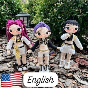 Puede incluir: Tres muñecas de ganchillo hechas a mano con cabello rosa, morado y negro. Cada muñeca viste un atuendo único con detalles dorados, incluyendo chaquetas, faldas y pantalones cortos. Las muñecas están sobre un lecho de astillas de madera.