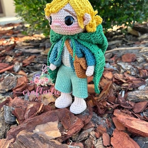 Puede incluir: Muñeca de ganchillo hecha a mano que representa a un personaje con pelo amarillo, una capa verde y una bandolera marrón. La muñeca lleva una camisa azul, pantalones cortos verdes y zapatos blancos. La muñeca está sobre mantillo marrón.