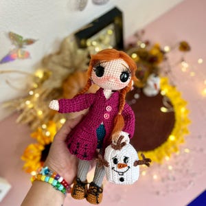 以下が含まれることがあります： 手作りの編み人形。赤い三つ編みの髪で、マゼンタ色のコートを着て、白い雪だるまのバッグを持っています。人形は大きな黒い目を持ち、茶色の靴を履いています。人形の高さは約18cmです。