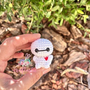 May include: A small, white crocheted figure with black eyes and a red heart. The figure is held in a hand against a background of green foliage. The figure is a handmade item.