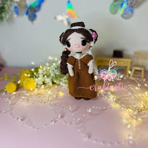 May include: A brown crocheted doll with a white dress and brown hair. The doll has a pink flower on her head and is wearing a necklace. The doll is standing on a pink surface with a string of lights in the background.