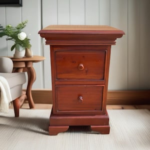 May include: A reddish-brown wooden bedside cabinet featuring two stacked drawers with small round knobs. The piece has a flat rectangular top with moulded edges and a sturdy base with cutout feet, showcasing a natural wood grain finish throughout.