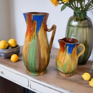 May include: Two ceramic pitchers featuring a drip glaze finish in vibrant shades of blue, orange, yellow, and green. Both pitchers have a curved handle and a pouring spout, with the larger pitcher standing taller than the smaller one.