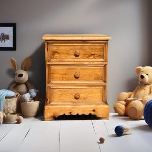 May include: A light brown wooden chest of drawers with three horizontal drawers, each featuring a single round wooden knob. The chest has a rectangular top and a decorative scalloped base, showcasing a natural wood grain finish throughout.
