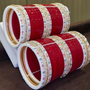 Può includere: Due set di braccialetti rossi e bianchi. I braccialetti rossi sono decorati con dettagli dorati e argentati e piccole gemme colorate. I braccialetti sono impilati, evidenziando il loro design intricato e i colori vivaci.