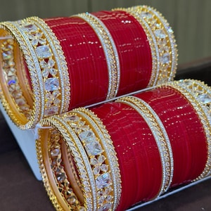 Può includere: Un set di braccialetti rossi e dorati. I braccialetti hanno un centro rosso brillante con bordi dorati. I bordi sono decorati con pietre preziose chiare, quadrate e a forma di fiore. I braccialetti sono impilati.