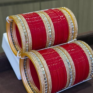 Può includere: Due set di braccialetti rossi con decorazioni dorate e argentate. I braccialetti hanno una finitura lucida e sono impilati. Gli accenti dorati sono sui bordi e al centro dei braccialetti.