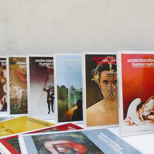 May include: A collection of vintage books titled "Understanding Human Behavior" with various cover illustrations. The covers feature images of people, abstract art, and text. Some books are stacked in front.