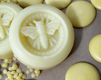 May include: A close-up of a round, white, beeswax candle with a bee design. The candle is surrounded by small, yellow beeswax pellets.