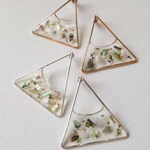 Puede incluir: Un par de pendientes de forma triangular con marcos de alambre dorado y plateado. Los pendientes están rellenos de resina transparente y contienen pequeños trozos de concha verde y blanca iridiscente.