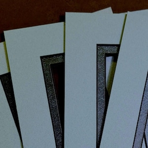 May include: Close-up of several off-white picture mats with black borders, slightly overlapping. The mats are arranged on a brown surface, creating a layered effect. The mats are designed for framing artwork or photographs.