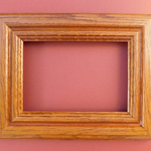 May include: A rectangular wooden picture frame with a warm, medium-brown finish. The frame has a ridged, layered design and a visible wood grain. The background is a solid, muted reddish-pink colour.