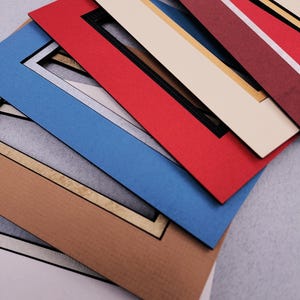 May include: A stack of rectangular paper picture frame mats in various colors including red, blue, tan, cream, and maroon. Each mat features a central rectangular cutout with a contrasting inner border, layered to show the different color options.