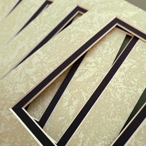 May include: A stack of beige picture mats with a textured surface and a black border. The mats have a rectangular opening in the center, designed for displaying photographs or artwork. The mats are stacked at an angle.