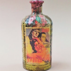 Può includere: Una bottiglia di vetro vintage decorata con ritagli di carta colorati e un poster di un film con una donna in un vestito rosso. Il poster recita "La Pervesa" e presenta il testo "Hollywood, Dolores regreso a Mexico..."