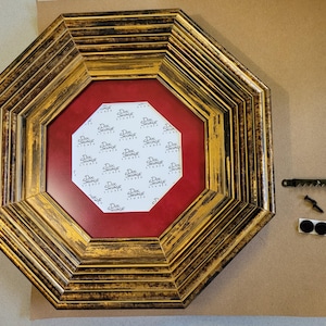 May include: An octagonal picture frame with a gold and black finish. The frame has a red interior and a white paper insert with the text "Doc Savage Studio". Includes hanging hardware and bumpers.