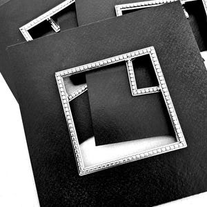 Op de afbeelding: Een verzameling zwarte vierkante kaarten met zilveren metalen frames. De frames hebben een vierkant ontwerp met een kleinere vierkante uitsparing. De frames zijn versierd met kleine, gelijkmatig verdeelde details.
