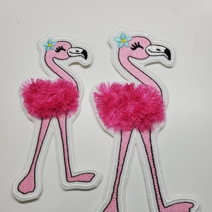 Op de afbeelding: Twee roze flamingo geborduurde patches met witte contouren. Elke flamingo heeft een blauwe bloem op zijn kop en een roze pluche rok.