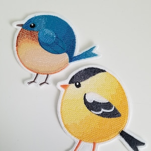 May include: Two embroidered iron-on patches of birds. One is a bluebird with a brown breast and the other is a yellow bird with a black head and wings.