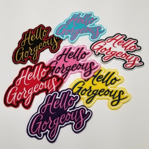 Può includere: Un set di sette toppe da stirare con il testo "Hello Gorgeous" in diversi colori, tra cui rosa, rosso, blu, giallo e viola.