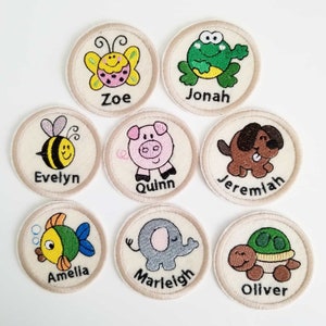 Könnte beinhalten: Acht gestickte Filzkreise mit Tiermotiven und Namen. Die Tiere sind ein Schmetterling, ein Frosch, eine Biene, ein Schwein, ein Hund, ein Fisch, ein Elefant und eine Schildkröte. Die Namen sind Zoe, Jonah, Evelyn, Quinn, Jeremiah, Amelia, Marleigh und Oliver.