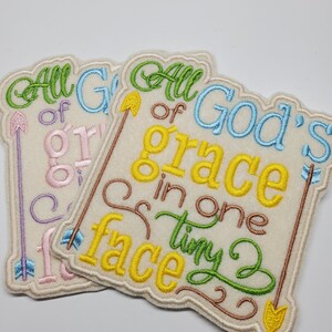 May include: Two embroidered patches with the text "All of God's grace in one tiny face". The patches are beige with colorful embroidery in green, yellow, blue, and pink.
