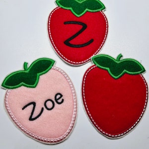 Könnte beinhalten: Drei Filz-Erdbeer-Patches, einer rot mit dem Buchstaben Z, einer rosa mit dem Namen Zoe und einer einfarbig rot.