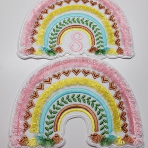 Puede incluir: Dos parches bordados de arcoíris con borde de flecos. Los arcoíris están superpuestos con bordados de color rosa, amarillo, azul y marrón. El arcoíris superior tiene una letra "S" rosa en el centro. Ambos arcoíris tienen hojas verdes y borlas marrones.