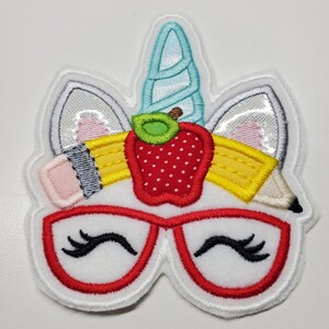 May include: A white, embroidered patch featuring a unicorn head with a light blue horn, silver ears, and red-rimmed glasses. A red apple and yellow pencils are above the glasses. The patch has a white border.