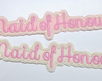 May include: Two white fabric patches with pink embroidered lettering that reads "Maid of Honour".